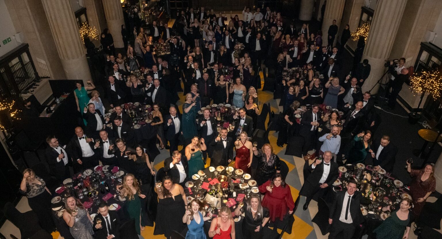 A large group of people in formal attire gathered in a grand hall with high ceilings and columns, seated at round tables with floral centerpieces. Many are standing, raising glasses, or posing for the camera.