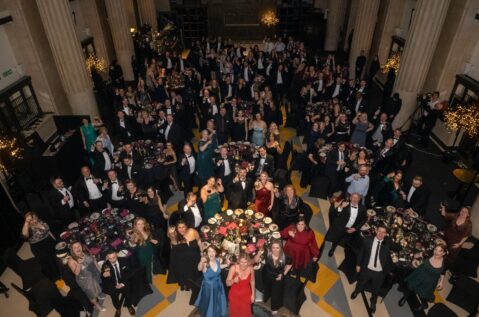 A large group of people in formal attire gathered in a grand hall with high ceilings and columns, seated at round tables with floral centerpieces. Many are standing, raising glasses, or posing for the camera.
