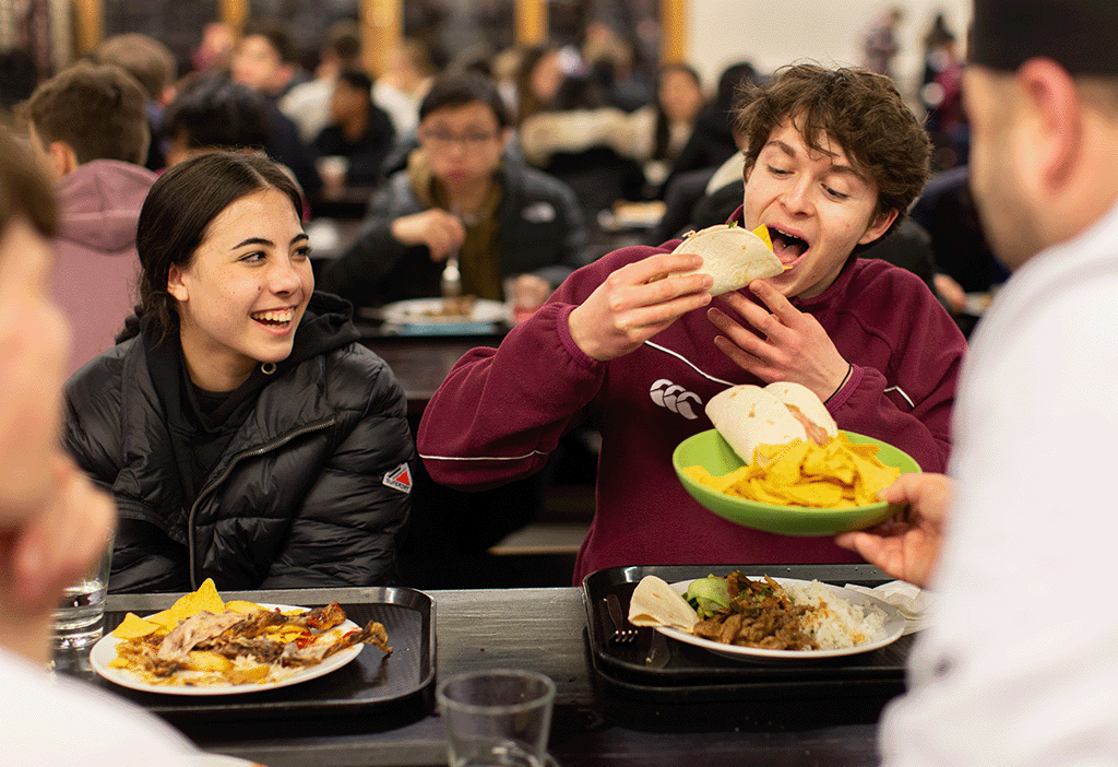 Bespoke catering services. Two pupils sit at a table in a busy dining hall. One is eating a taco, both have trays of food.