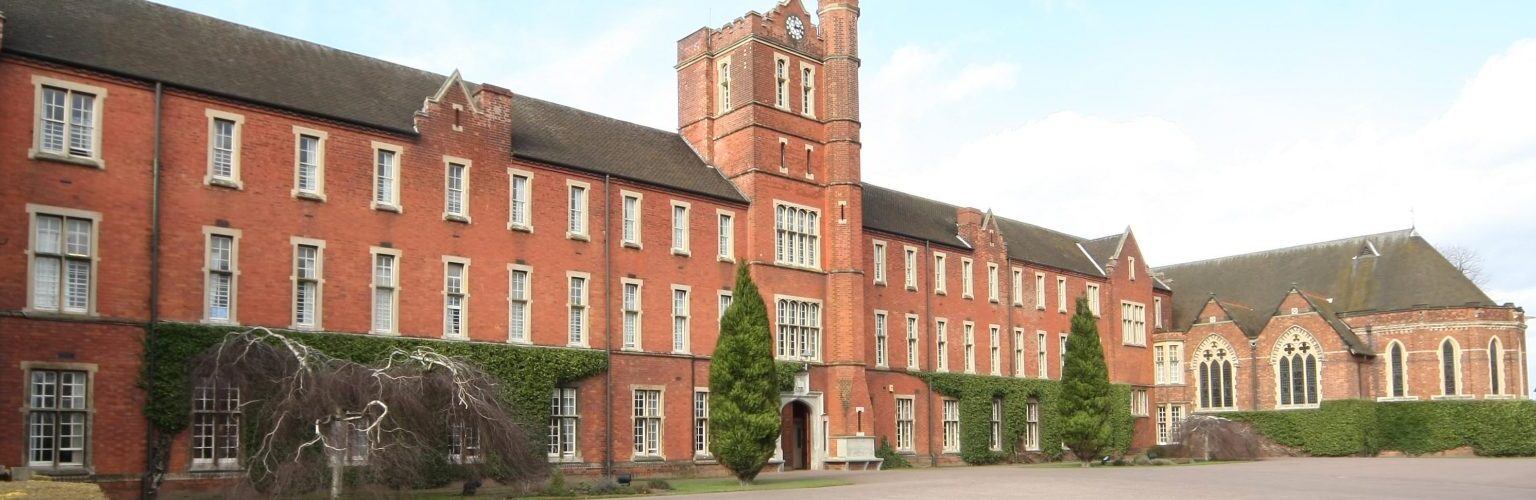 A picture of Trent College;A historic red-brick building with a clock tower, surrounded by hedges and trees. The sky is clear with some clouds.