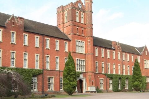 A picture of Trent College;A historic red-brick building with a clock tower, surrounded by hedges and trees. The sky is clear with some clouds.