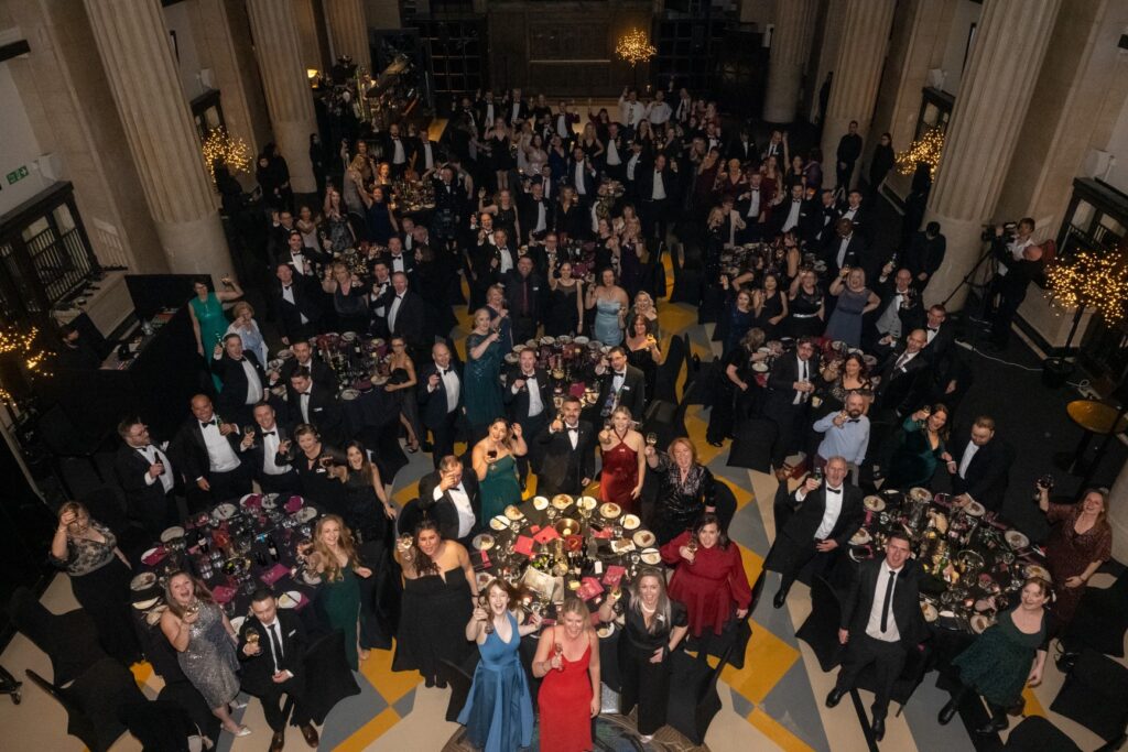A large group of people in formal attire gathered in a grand hall with high ceilings and columns, seated at round tables with floral centerpieces. Many are standing, raising glasses, or posing for the camera.