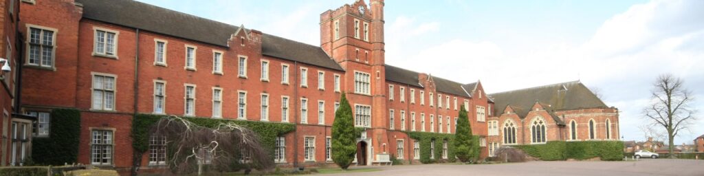 A picture of Trent College;A historic red-brick building with a clock tower, surrounded by hedges and trees. The sky is clear with some clouds.